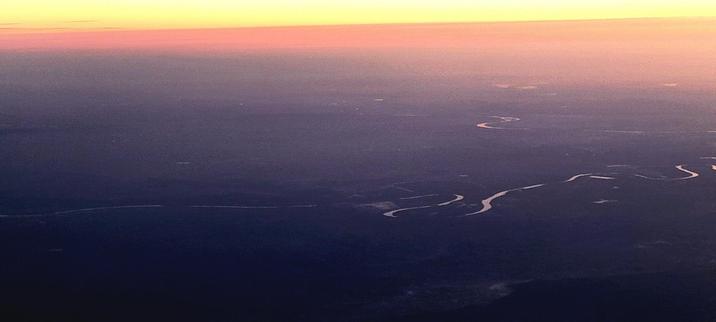 View of landscape and horizon from inside an aircraft. Purple, pink, and yellow skies. Purple landscape with a windy river.
