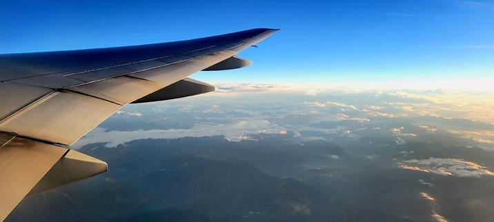 Part of the right wing of a commercial jet aircraft seen from inside the aircraft. Soft white light from a just risen sun falls on the wing. Blue skies with white fluffy clouds. The back/top of a mountain range visible below.