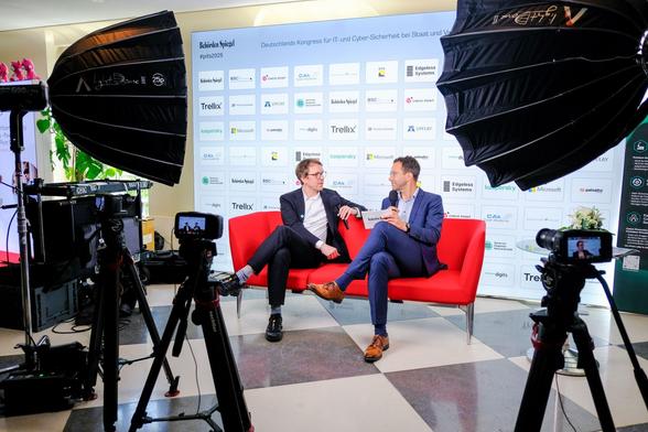 Leonhard Kugler und Guido Gehrt sitzen auf einem roten Sofa. Beide halten Mikrofone in der Hand. Um das Sofa herum sind Lichter und Kameras aufgebaut.