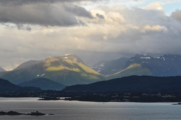 A photo of a body of water with mountains in the distance. The mountains are green with small amounts of snow on them. Light is coming through the cloudy sky onto the mountains and casting shadows on the lower closer land.
