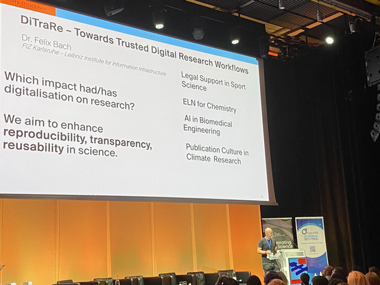 Felix Bach from FIZ Karlsruhe presenting the DiTraRe poster at the Open Science Fair at CERN, Geneva, Switzerland. A poster teaser slide is presented on a large screen behind Felix. Its headline says: DiTraRe - Towards Digital Research Workflows. Below it states: "WHich impact has/had digitalisation on research?", and "We aim to enhance preproducibility, transparency, reusability in science."