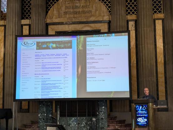 A white male speaker behind a speaker's desk with the conference logo displayed at the desk front, and a large screen embedded in the walls of an old building behind him. The screen shows an overview of various talks scheduled for the afternoon.