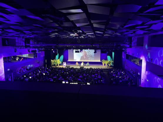 Photo of a large lecture hall with a big stage on the front viewed from a seat on the second balcony. The illumination is mostly purple. On the stage, three women are seated in armchairs, a fourth one is giving a speech remotely while being displayed on a large screen above the others. Potted plants frame the stage on either side, the rows of seats in front of the stage are completely filled with people.