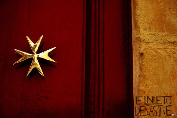 Fotografía de una cruz de malta con fondo rojo y amarillo de una fachada de una casa
