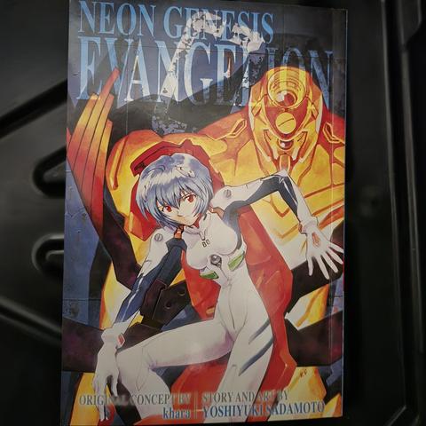 Photograph of Neon Genesis Evangelion manga (omnibus 2) placed against a black colored surface. The title is in the upper portion of the cover. Beneath that in the background is a yellowish robotic looking creature (Eva Unit 02) appearing stoic and immobile. In the foreground is a light blue haired teenage girl in a tight, white suit sitting in some kind of pilot's chamber.