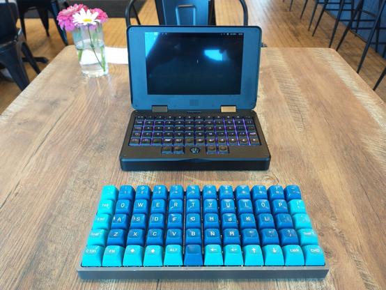 An MNT Pocket Reform (a surprisingly tiny laptop with a 5x12 ortholinear mechanical keyboard) sitting on a wooden table behind a Preonic ortholinear mechanical keyboard of the same physical configuration.  The labels on the key caps are mostly equivalent.  The Preonic has blue and cyan key caps in the Data General Dasher color scheme.  There are fresh pink and white flowers in a glass of water behind the laptop.