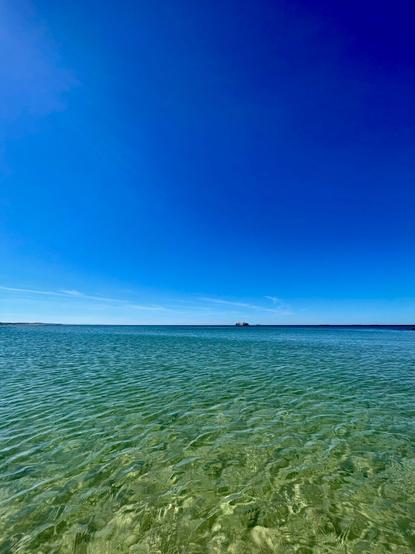 A clear, calm sea with gentle ripples, reflecting a bright blue sky. A distant island is visible on the horizon.