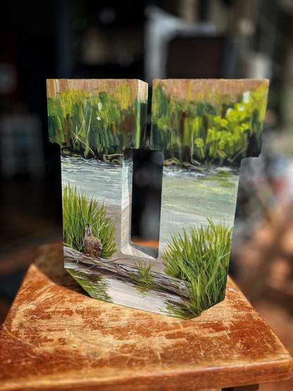 A blocky wooden letter “U” painted with a marsh scene, showing tall green reeds along the water’s edge. A small duck stands on a log in the foreground surrounded by grasses, with the water and reeds continuing in the background.