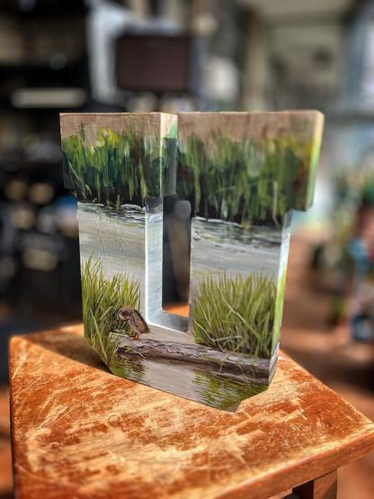 The reverse side of the same painted letter “U,” depicting the continuation of the marsh scene. Another duck is perched preening on the same log, partially hidden among the grasses, with water and dense reeds extending across the background.