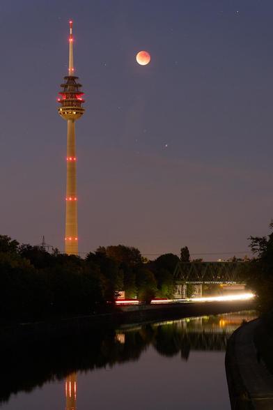 Nachtaufnahme des Nürnberger Fernmeldeturms während der totalen Mondfinsternis: im unteren Drittel des Bilds ein breiter Kanal, in dem sich der unbewölkte Himmel spiegelt, und darüber die durch Langzeitbelichtung verwischten Lichter von Autos auf einer Uferstraße. Am Turm leuchten rote Positionslichter auf mehreren Ebenen, und neben dem Turm in der Bildmitte leuchtet der rötliche Vollmond am Himmel.