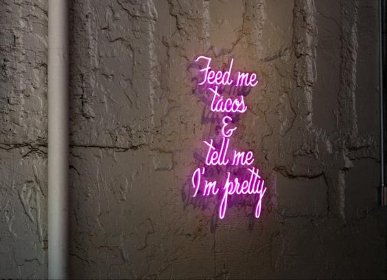 A neon word sign on a rustic Mexican restaurant wall that says, "Feed me tacos & tell me I'm pretty.