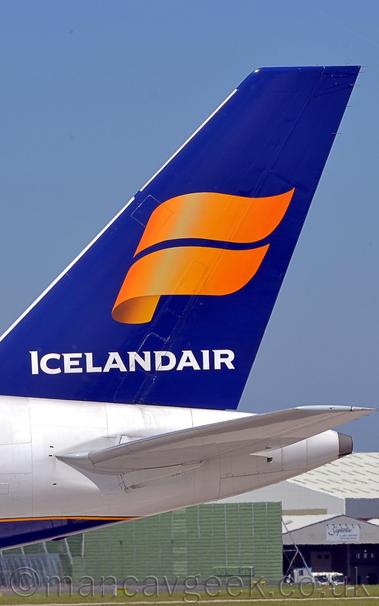 Close-up of the rear fuselage and tail if a jet airliner facing to the left.
The plane has a mostly white body, with a dark blue belly and a yellow pinstripe.
The plane's tail is the same dark blue, with a yellow, twisted double ribbon, and white "IcelandAir" text at the bottom.
Under the plane's tail, green grass in the background leads up to a boxy, green metal structure in the distance, with a pair of grey hangars beyond.
Pale blue sky fills the rest of the frame.