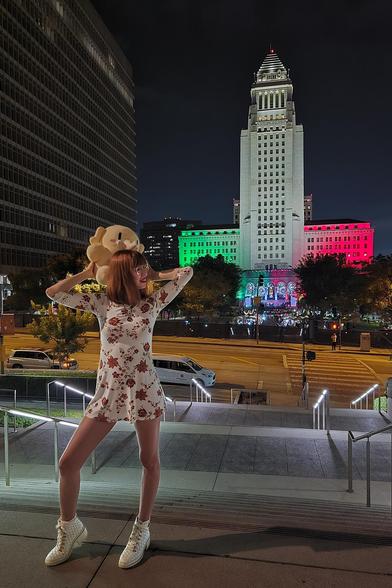 Foto de Lety y juguete de peluche Creature Mastodon parados frente al Ayuntamiento de Los Ángeles, iluminado con los colores de la bandera de México durante una noche oscura. De arriba a abajo, Lety viste un vestido corto blanco de manga larga con estampado floral rojo y botas blancas de cuero. Está sobre unas escaleras a dos calles del ayuntamiento, con el cuerpo de frente a la cámara, la pierna derecha extendida y la rodilla derecha doblada. Lety sostiene al Creature con ambas manos cargándolo en la espalda como caballito detrás de su cabeza, mientras ambos voltean hacia el edificio iluminado.