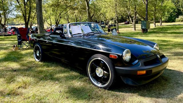 Photograph of a black 1976 MGB facing right in a park with green trees and grass.