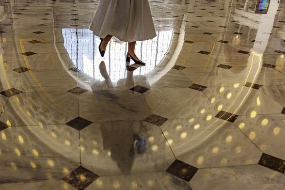 A woman in a white flounce dress and black hills is crossing Union Station in Washington DC only the bottom part of her body with the moving dress and feet and her shadow reflection on the marble floor are visible. The stainglass window and the archs of the ceiling reflected on the floor shape a frame around her body.