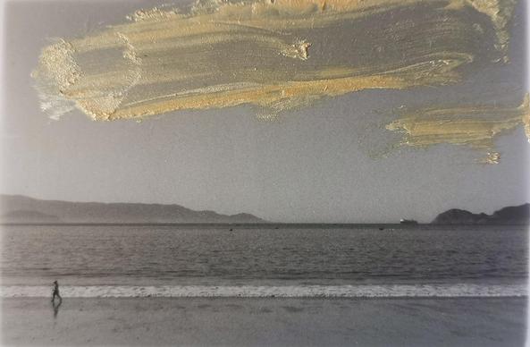 Playa al atardecer. Foto en blanco y negro. Casi al limite a la izquierda, una una figura solitaria camina junto a la orilla, bajo un cielo intervenido con pinceladas doradas y texturizadas. El mar y las montañas difuminadas completan una escena de quietud y melancolía visual.