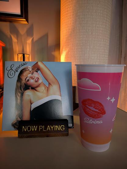 'Espresso' 7" vinyl single in a "Now Playing" stand, a candle in the background, and a pink Sabrina cup with a lipstick print on it from Dunkin