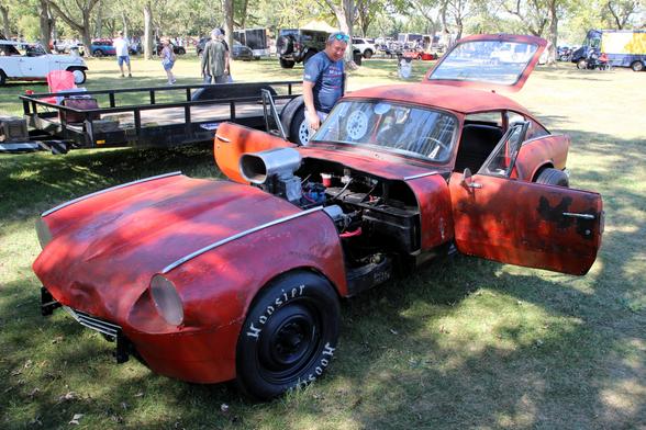 Photograph of an old red Triumph GT6 body with an engine scoop sticking out. The car is on grass in a park.