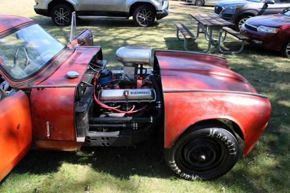 Photograph of an Oldsmobile 440 V8 engine with an engine scoop in an old red Triumph GT6 body. The car is on grass in a park.