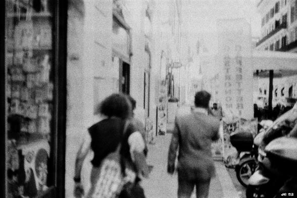 Etwas unscharfes Schwarzweißfoto, das eine Straßenszene in Rom zeigt. Links ein Ladenfenster, in Bildmitte die Rücken von drei Personen, rechts geparkte Motorroller, im Hintergrund hohe Gebäude. Olympus 35SP von 1969 auf Kentmere 100.