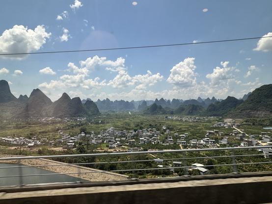 Lots and lots of small narrow hills repeating off into the distance, with some reflections from the train window I’m looking through. A few clouds in the blue sky beyond, and in front some low-density housing among the green.