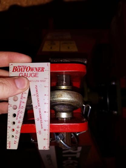 A small credit card sized combination ruler/gauge with holes for sticking screws through and various useful labels on both sides. It's being held to a tractor implement to measure the length of a pin.