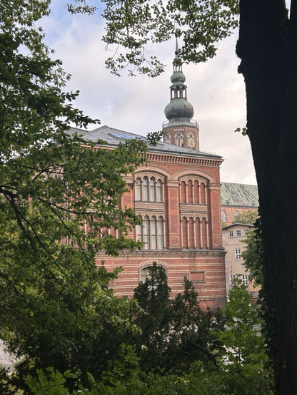 Blick auf altes Unigebäude. Ein Ziegelgebäudr mit hohen Fenstern. Dahinter der gotische Turm einer der Kirchen in Greifswald.