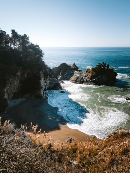 A coastal view with waves crashing on sandy shores, surrounded by rocky cliffs and pine trees under a clear blue sky.