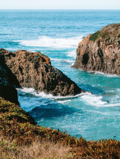 Rocky formations dividing turquoise waters with waves gently rolling in, surrounded by lush greenery and a clear sky.