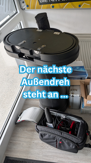 Ein runder, schwarzer Transportkoffer steht neben einer geöffneten, grauen Kameratasche. In der Tasche sind ein schwarzes Mikrofon, rote Kabel und weiteres technisches Equipment zu sehen. Im Hintergrund sind Kisten und Bücher gestapelt.