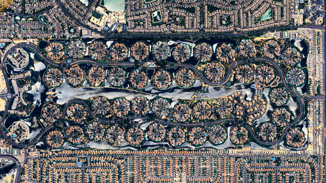A satellite picture of a residential area featuring circular clusters of houses arranged in a winding pattern, surrounded by water canals. The design resembles islands connected by internal roads, with traditional grid-like neighborhoods and larger buildings surrounding the central serpentine layout.