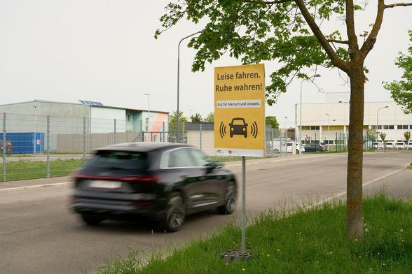 Ein Auto fährt auf einer Straße vorbei. Im Vordergrund steht ein gelbes Schild mit dem Text 'Leise fahren. Ruhe wahren!'. Im Hintergrund sind Gebäude und ein Baum sichtbar. Foto: Andreas Fuchs, AIT