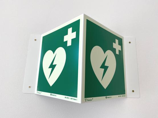 Dunkelgrün-weißes Hinweisschild an der weißen Wand, direkt über dem Defi