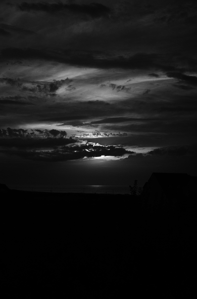 A black and white photography depicting a dark sky with a view on the sea. The sun is just behind a cloud.