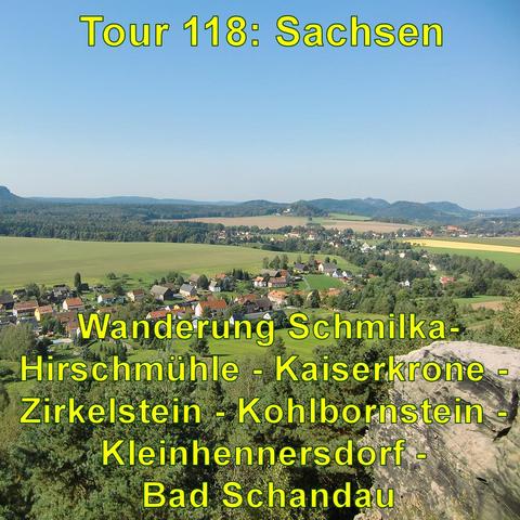 Blick von einem erhöhten Standpunkt auf eine ländliche Landschaft: Im Vordergrund ein Dorf mit mehreren Häusern, umgeben von Bäumen. Dahinter erstrecken sich Felder und weitere kleine Waldstücke. In der Ferne sind sanfte Hügel zu erkennen, die den Horizont bilden. Der Himmel ist klar und blau.