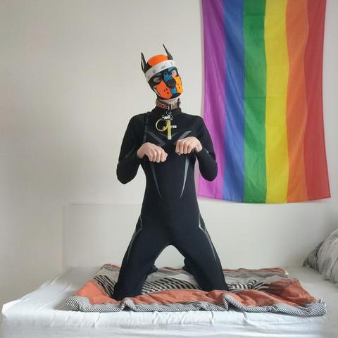 Aki kneeling on a bed in a black wetsuit in front of a pride flag