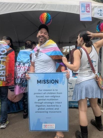 Intact global board member, Che, to the left holding Intact Global's mission statement poster.   Alliana, GAC founder, standing facing away from the camera showing off the GALDEF logo on the back of her tank top.  Both in front of the GALDEF Table at Portland pride 2025.