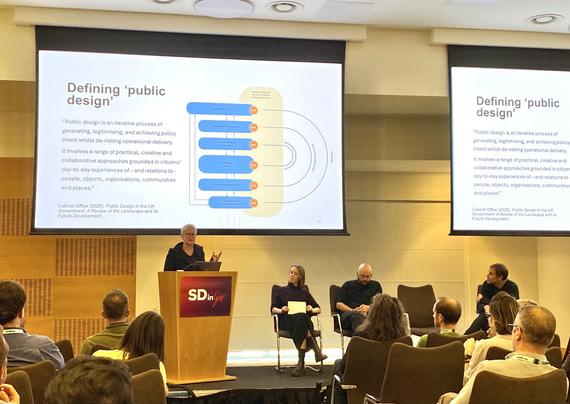 3 people on a conference stage sitting in front of an audience, 1 woman with grey hair standing at a lectern speaking, a projected slide on top of them says “defining public design — “Public design is an iterative process of generating, legitimising, and achieving policy intent whilst de-risking operational delivery.
It involves a range of practical, creative and collaborative approaches grounded in citizens' day-to-day experiences of - and relations to - people, objects, organisations, communities and places.”; next to it, a version of a policy cycle