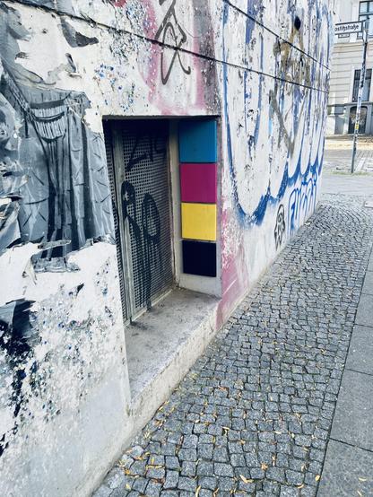 CMYK: Schrägansicht auf ein Kellerfenster an einem alten Mietshaus. Jede Menge Graffiti an den Wänden aber auch vier quadratische Plättchen in den Farben CMYK (Himmelblau, Magenta-Rot, Gelb und Schwarz).