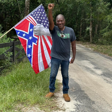 Dr. Marvin Dunn, an 85 year old Black man, is standing in a rural road in Florida with a determined smile but also a raised fist. He's wearing a dark grey T shirt, jeans, and tan colored boots. He is standing next to a US flag that has been made to look like a hand is pulling it away to reveal a Confederate flag underneath, a symbol of Florida Republicans' hypocrisy.
