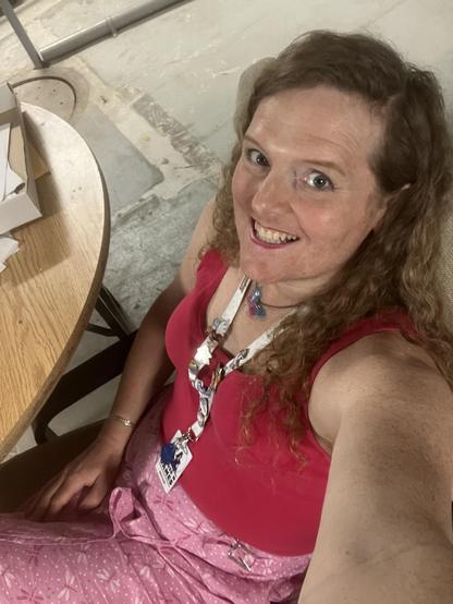 miriam, a white trans woman with long curly dirty blonde hair, in a raspberry tank top and pink skirt decorated in dragonflies. she wears a name badge on a lanyard decorated with various pride flag ename pins