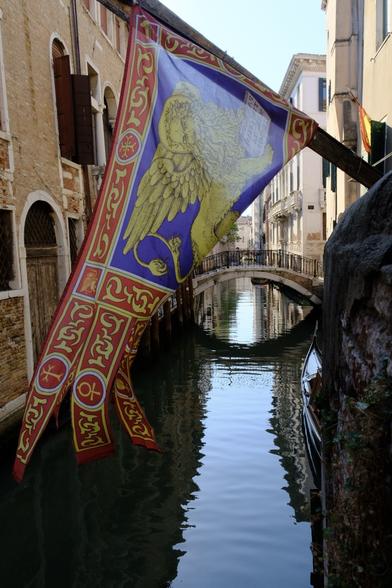 A view of a canal in Venice. In the foreground is the flag of Venice, hanging from an oblique, almost horizontal pole, which is attached to a wall on the right edge of the scene. The flag is swayed by the wind and lifts just enough to open the view to the canal behind it, passing under a pittoresque bridge into the distance. In both sides of the canal there are typical Venetian palazzos.