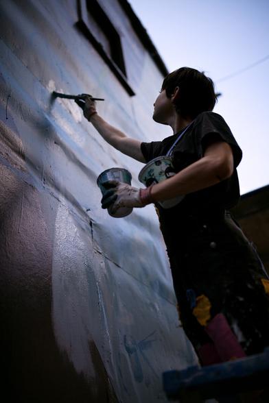 A low angle shot of a female artist holding two cups of paint and a brush, there is a high contrast and backlight that gives an aery impression.