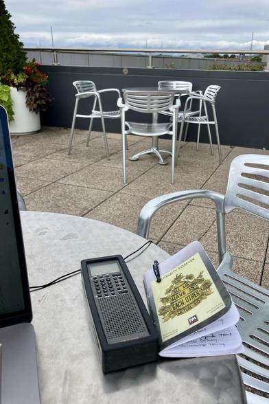 A gray radio on a silver table, next to a small paper notebook. The sky is in the background, behind another table and silver chairs.