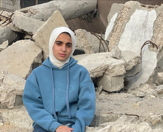 Foto de una mujer joven con una sudadera azul y un pañuelo blanco en la cabeza, con gesto serio mirando a cámara. Detrás de ella solo ruinas de un edificio