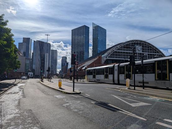 A wide road with tramways passes a station with tall buildings in the distance.