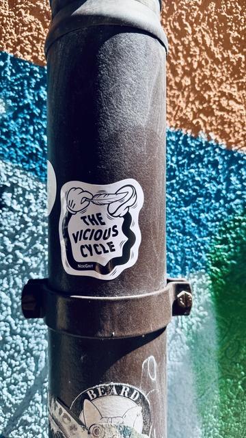 Sticker on an water pipe representing a cartoonish hand linked to foot by a black limb and is ticking the foot with a feather. The text on the sticker says «  The Vicious Cycle » by NoeGrit
