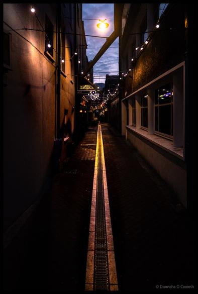 Atmospheric night photograph of narrow Carey's Lane in Cork showing illuminated string lights overhead, wet cobblestones with yellow central drainage channel, a restaurant sign, and moody twilight sky visible between buildings.