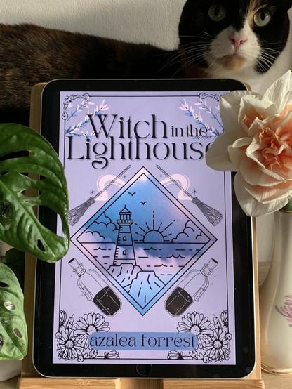Witch in the Lighthouse by Azalea Forrest, the cover displayed on a tablet with a calico cat behind it, surrounded by flowers and plants. Image taken by Karen Lykkebo (thank you!)