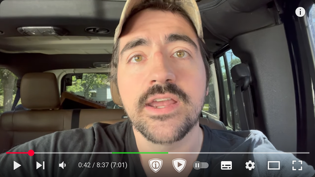 a white man in a baseball cap, talking in his car